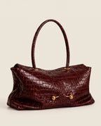 A burgundy crocodile-embossed leather handbag with dual top handles and gold-tone hardware, shown against a solid light beige background.