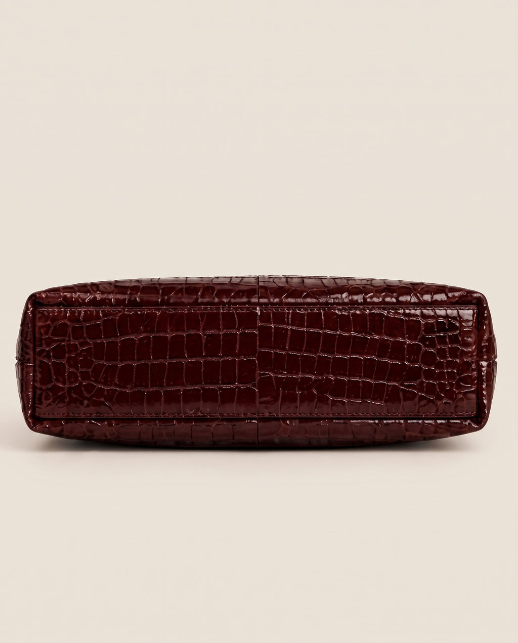 A dark burgundy crocodile-embossed leather clutch or pouch, shown from the bottom side against a solid light beige background.