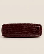 A dark burgundy crocodile-embossed leather clutch or pouch, shown from the bottom side against a solid light beige background.