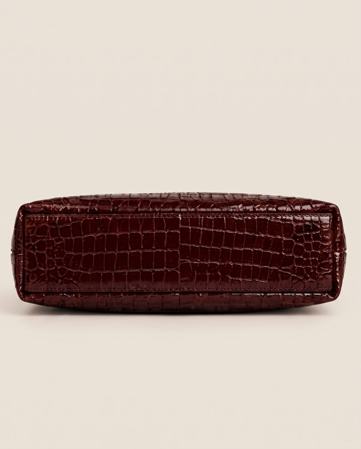 A dark burgundy crocodile-embossed leather clutch or pouch, shown from the bottom side against a solid light beige background.