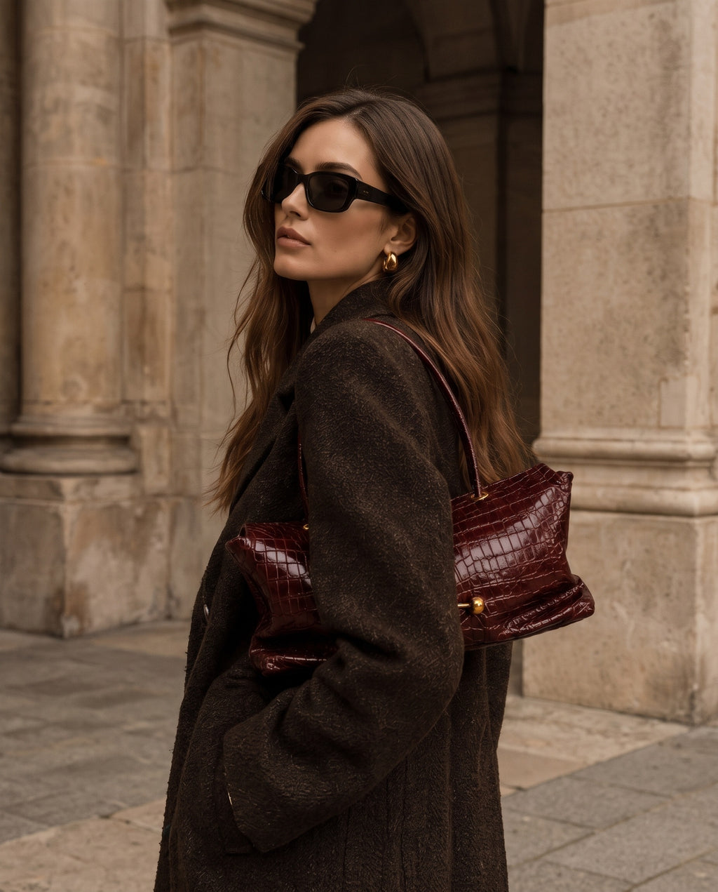 A woman in a dark brown textured coat and black sunglasses carries a dark burgundy crocodile-embossed leather shoulder bag, standing in front of a stone building.