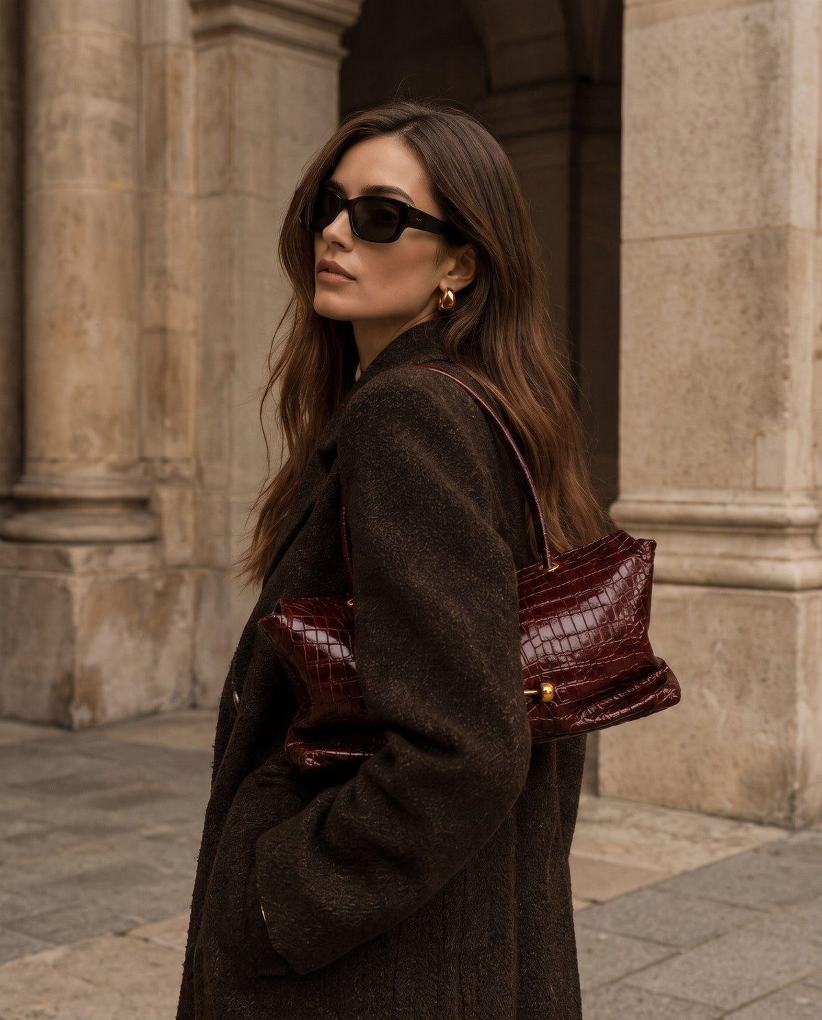 A woman in a dark brown textured coat and black sunglasses carries a dark burgundy crocodile-embossed leather shoulder bag, standing in front of a stone building.