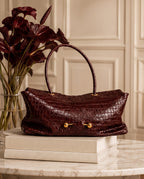 A dark burgundy crocodile-embossed leather handbag with gold-tone hardware, displayed on a stack of books on a marble table next to a vase of red flowers.