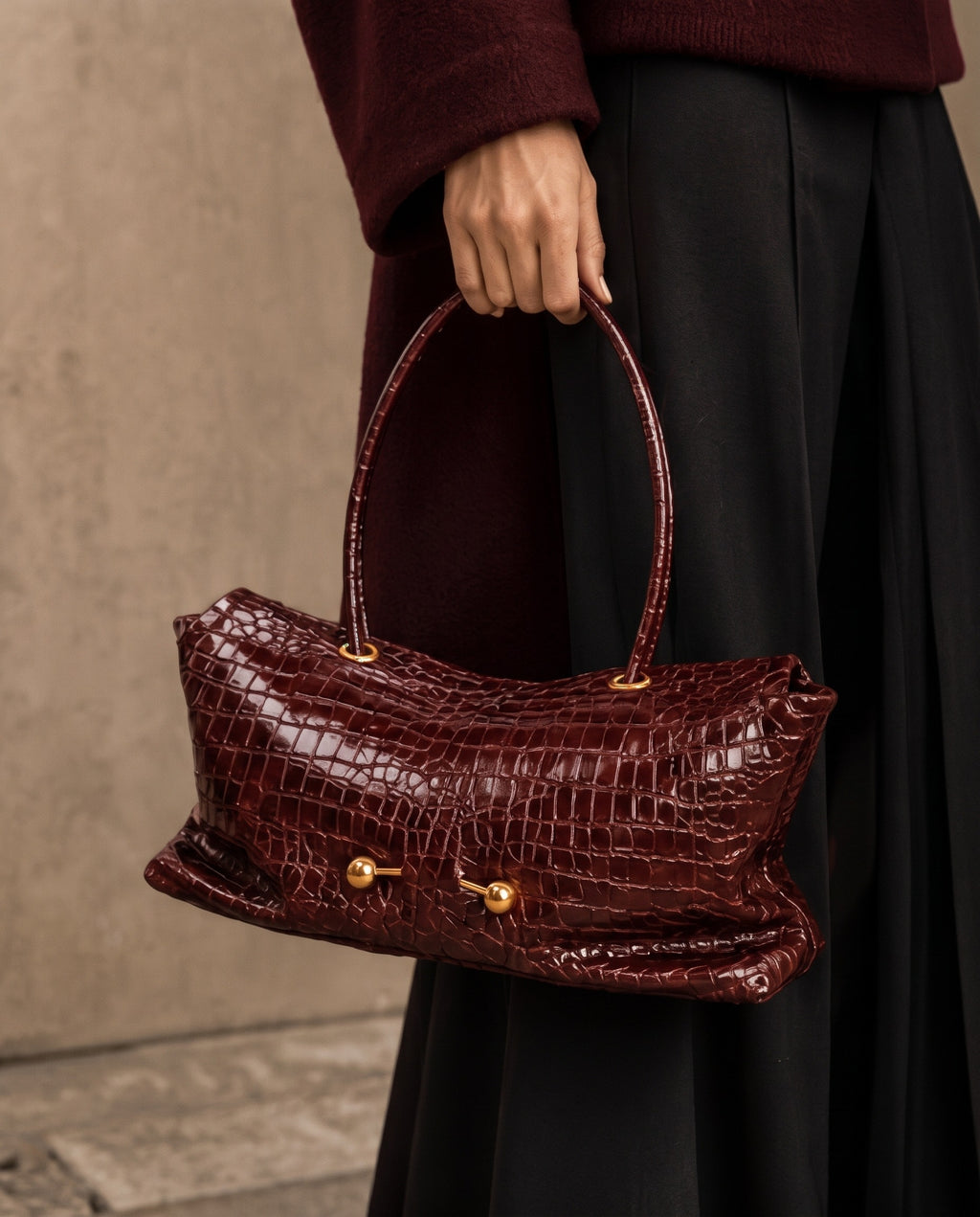 A person in a burgundy sweater and black skirt holds a glossy dark burgundy crocodile-embossed handbag with gold-tone hardware against a textured beige wall.