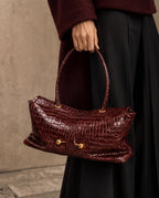 A person in a burgundy sweater and black skirt holds a glossy dark burgundy crocodile-embossed handbag with gold-tone hardware against a textured beige wall.
