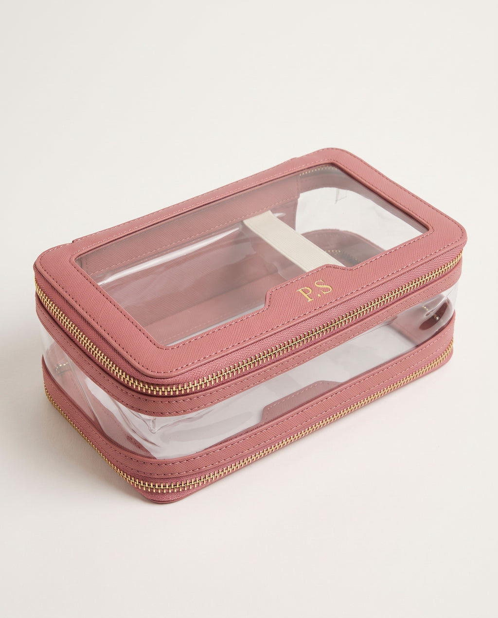 A rectangular makeup organizer case with a dusty pink faux-leather exterior, clear plastic window top, and gold zipper. The initials 'PS' are monogrammed in gold on the front window.