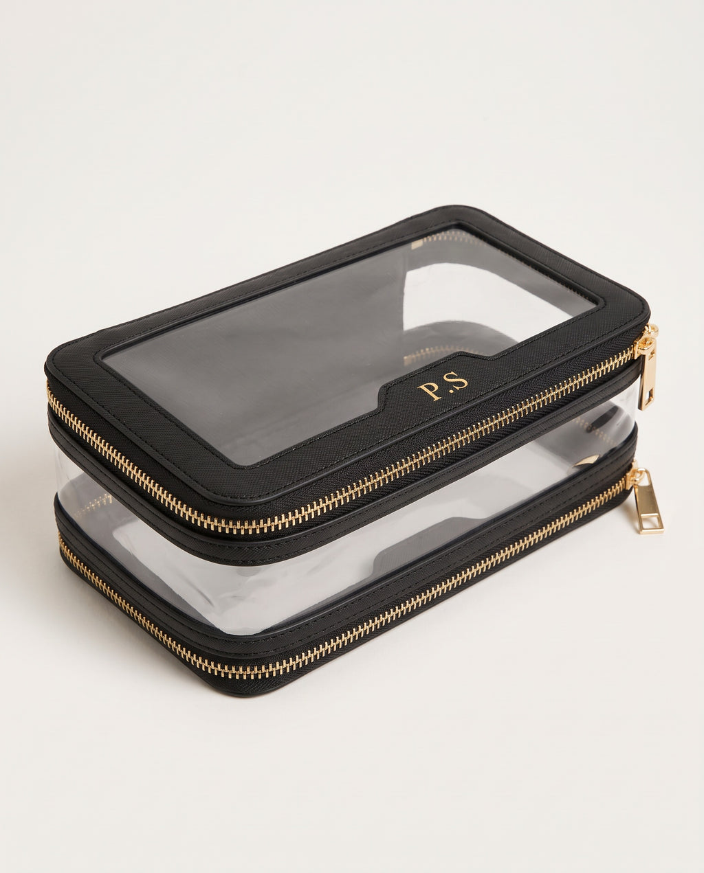 A black faux-leather zippered pouch with a clear window on top and a second clear zippered layer underneath, featuring gold zippers and a gold monogram 'PS' on the clear window.