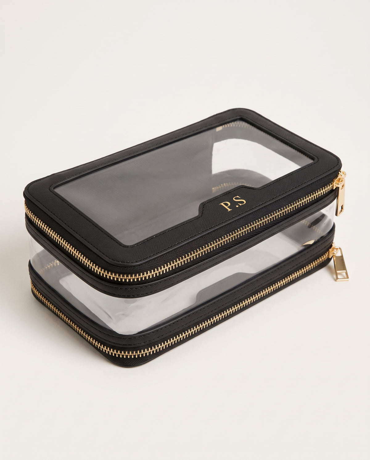 A black faux-leather zippered pouch with a clear window on top and a second clear zippered layer underneath, featuring gold zippers and a gold monogram 'PS' on the clear window.