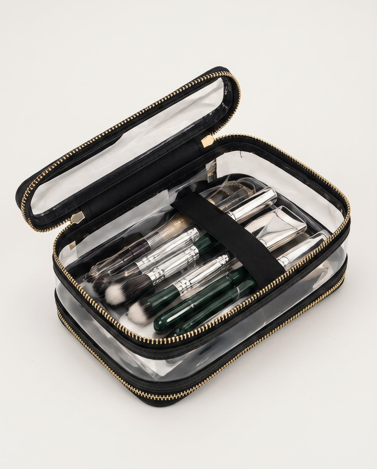 A clear zippered makeup bag with black trim and a black elastic band, shown open on a light gray background. Inside are several makeup brushes with silver and dark green handles.