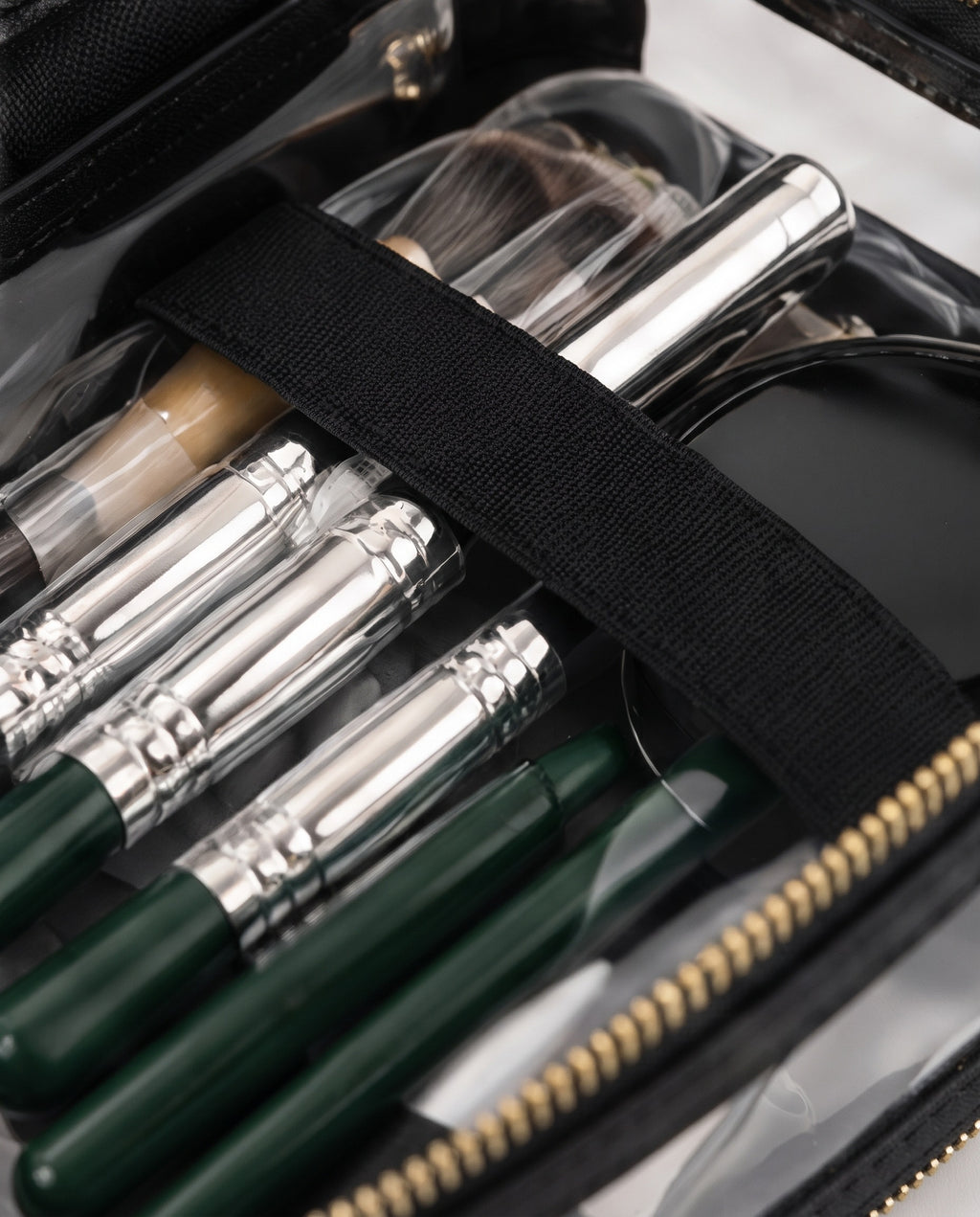 Close-up of a makeup brush and tool organizer inside a clear zip case. The organizer contains several makeup brushes with dark green handles and silver ferrules, along with other cosmetic tools secured by a black elastic band.