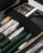 Close-up of a makeup brush and tool organizer inside a clear zip case. The organizer contains several makeup brushes with dark green handles and silver ferrules, along with other cosmetic tools secured by a black elastic band.