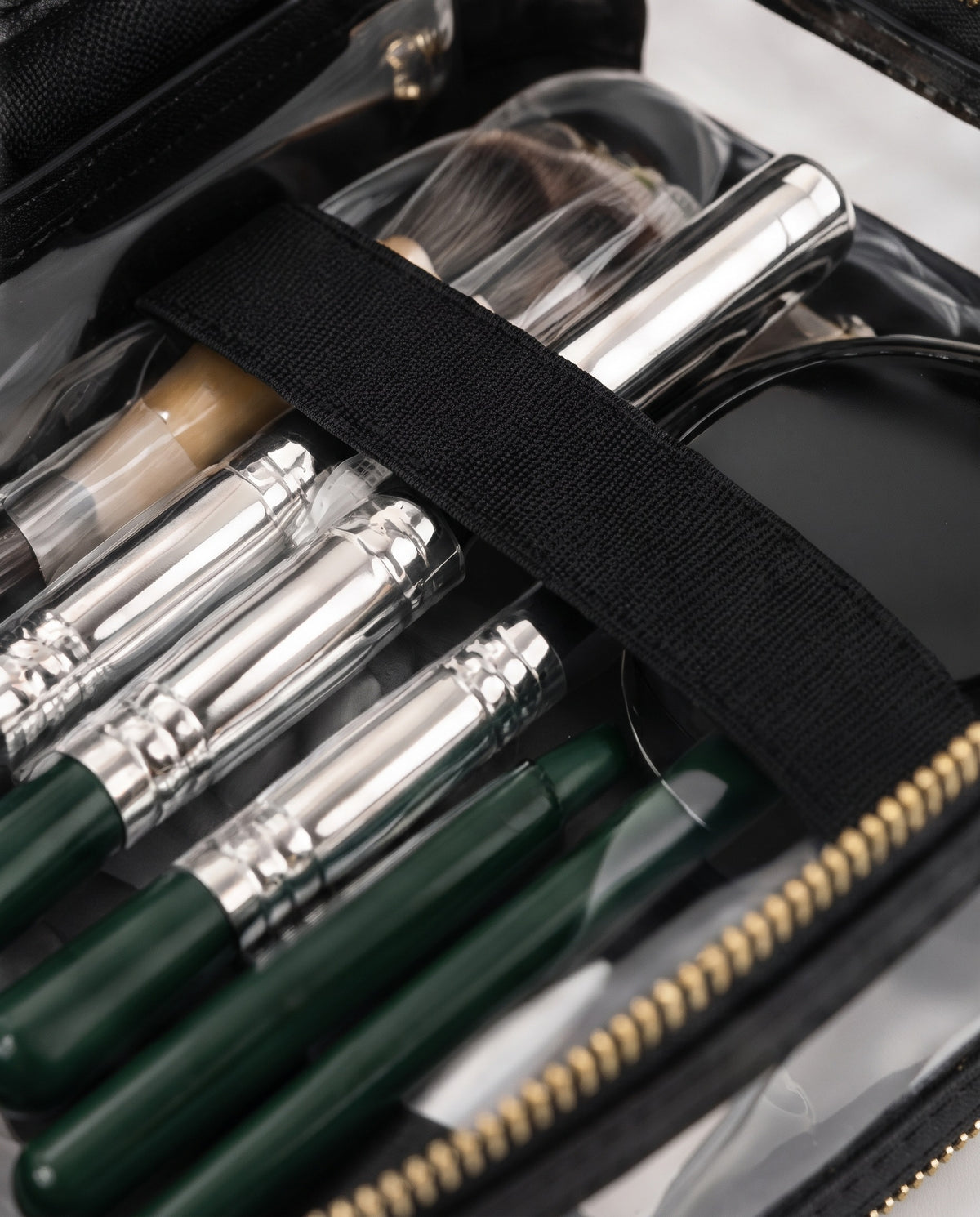 Close-up of a makeup brush and tool organizer inside a clear zip case. The organizer contains several makeup brushes with dark green handles and silver ferrules, along with other cosmetic tools secured by a black elastic band.