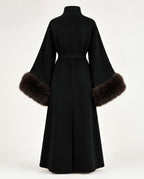 A black long-sleeved wrap coat with a high collar and a matching waist belt, featuring dark brown fur trim at the wide sleeves, shown from the back on a white studio background.