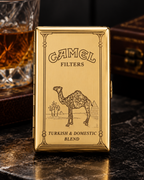 A gold Camel Filters tin for Turkish and Domestic Blend tobacco, featuring an engraved camel illustration, displayed on a dark marble surface with a glass of amber liquid in the background.