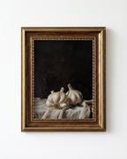 A framed still life painting of garlic bulbs resting on draped white fabric against a dark background, enclosed in an ornate gold-toned frame and mounted on a white wall.