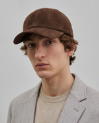 A close-up of a man wearing a brown suede baseball cap, a light gray blazer, and a beige knit sweater against a plain light gray background.