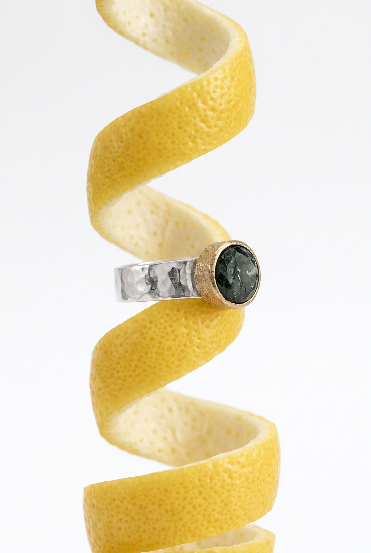 A silver and gold ring with a dark green cabochon stone, displayed horizontally against a spiral lemon peel on a white background.