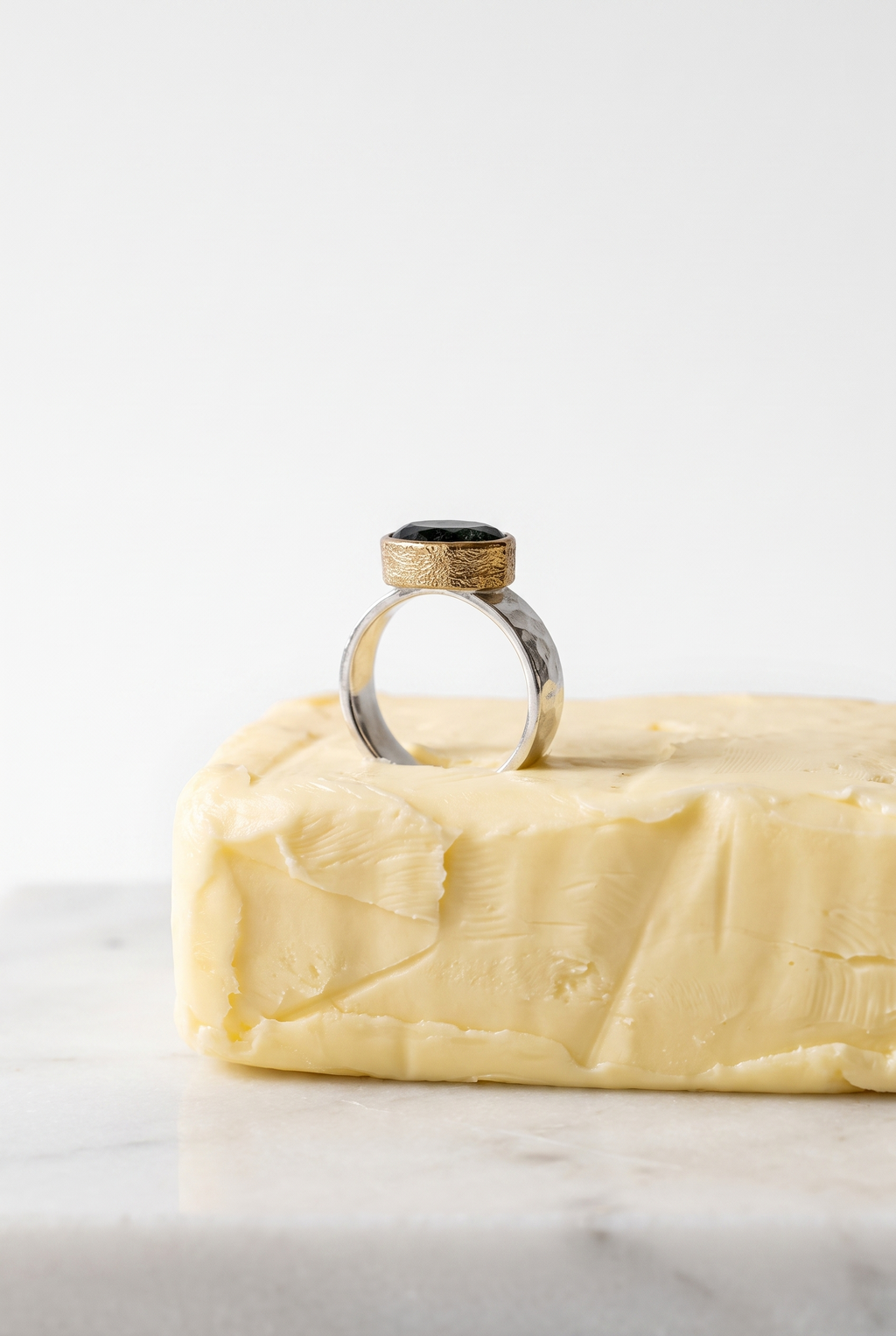 A silver ring with a textured gold band and a black oval stone, displayed on top of a block of butter on a white marble surface against a white background.