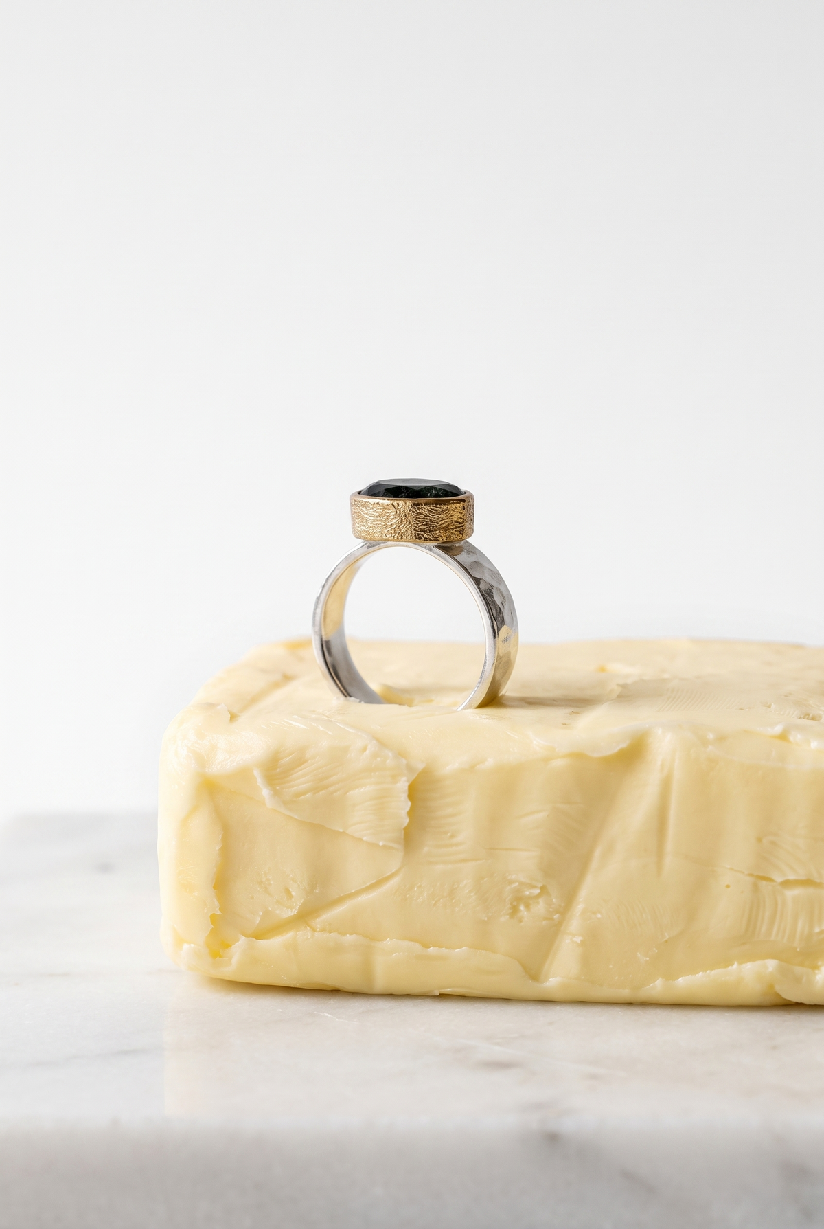 A silver ring with a textured gold band and a black oval stone, displayed on top of a block of butter on a white marble surface against a white background.