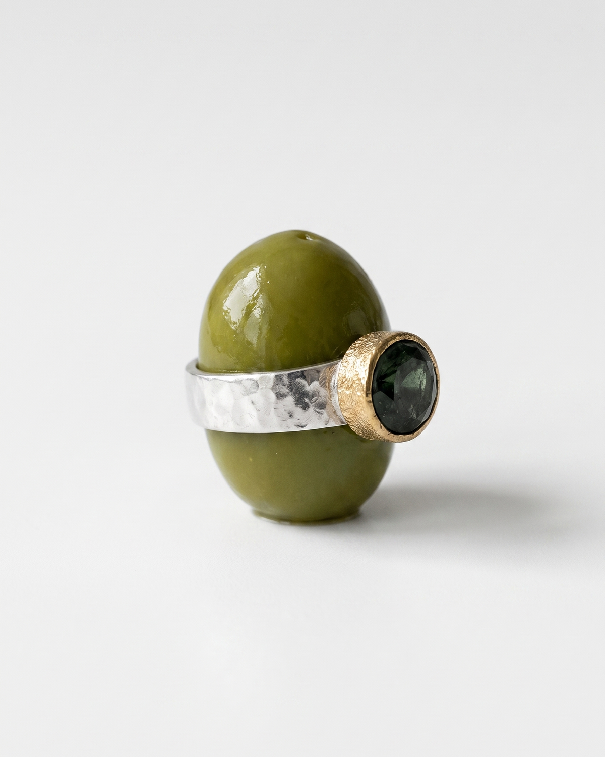 A ring displayed on an olive green object against a light gray studio background. The ring has a hammered silver band with a textured gold-toned bezel holding a round dark green gemstone.