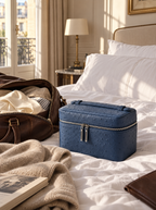 A blue textured leather cosmetic case sits on a neatly made bed in a sunlit bedroom, surrounded by travel props including an open brown leather bag, a beige knit blanket, and an open book.