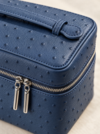A close-up of a navy blue textured leather cosmetic case with an embossed ostrich pattern, featuring a top handle and dual silver-tone zipper pulls.