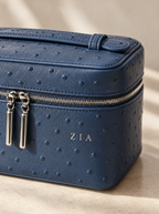 A close-up of a navy blue ostrich-embossed leather cosmetic case with silver zipper pulls and a gold monogram 'ZIA' on the front, set against a warm beige background.