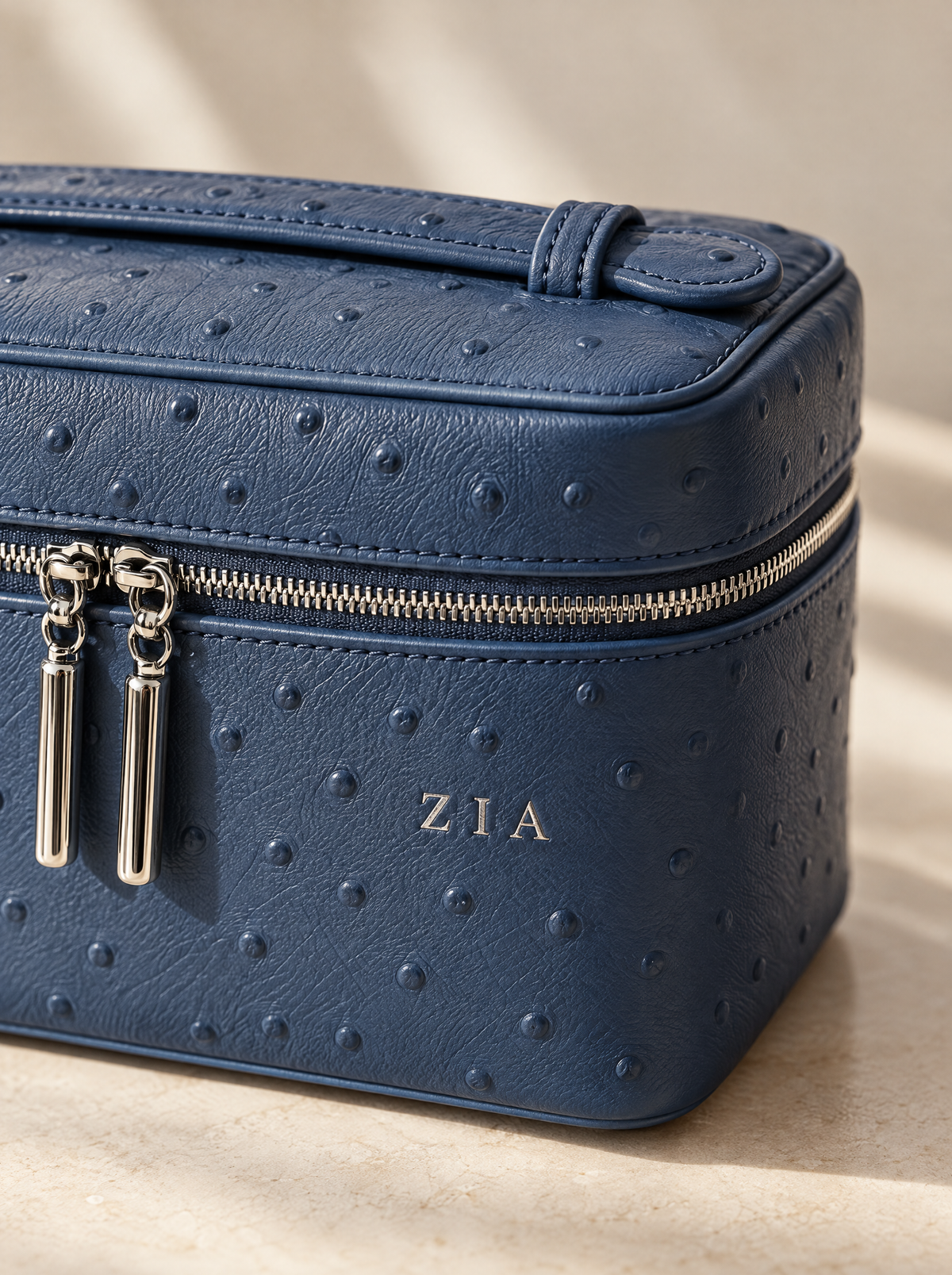 A close-up of a navy blue ostrich-embossed leather cosmetic case with silver zipper pulls and a gold monogram 'ZIA' on the front, set against a warm beige background.