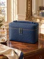 A navy blue ostrich-textured cosmetic case with silver zippers and a top handle, placed on a polished wooden vanity table in a luxurious room with an ornate gold mirror and a cream-colored scarf.