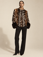 A model stands against a warm beige studio background wearing a brown and white spotted fur jacket with dark fur trim, paired with black trousers and black pointed heels.