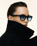 A close-up of a model wearing black cat-eye sunglasses with blue mirrored lenses and a high-collared black coat against a plain off-white background.