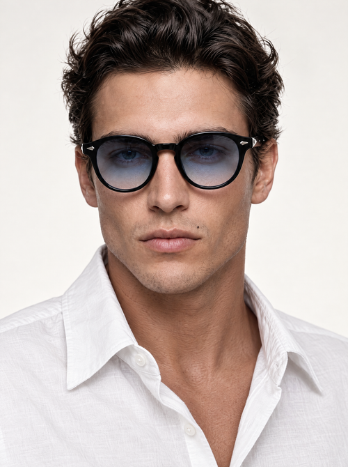 A close-up portrait of a man wearing black round-frame sunglasses with a subtle blue-gray gradient lens, paired with a white collared shirt against a clean off-white background.