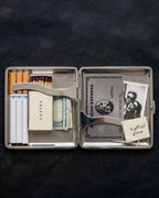 A silver metal cigarette case opened flat on a dark textured background, containing cigarettes, matches, a cardholder, a small photo, and a small note.