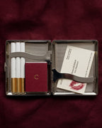 An open rectangular metal cigarette case on a dark burgundy fabric background, containing four cigarettes, a small burgundy pouch with a gold emblem, a metal lighter, and a printed card with a lipstick kiss mark.