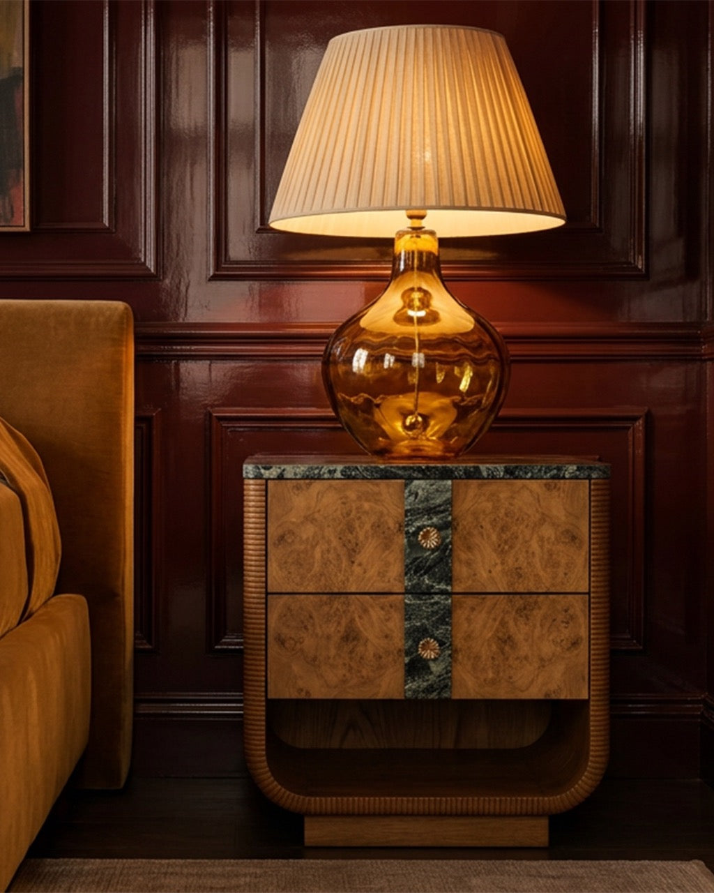 A decorative table lamp with a ribbed beige pleated shade and a warm amber glass base, placed on a carved wooden nightstand against a dark wood-paneled wall.