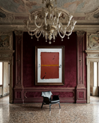 A framed abstract artwork with a terracotta red rectangle and a gold horizontal band, displayed on a deep burgundy wall in an ornate interior with a large chandelier, carved wood paneling, and a black leather chair below.