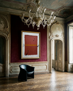 An abstract painting with a red and orange composition in a gold frame, displayed on a dark burgundy wall in an ornate classical room with a large crystal chandelier, a black leather armchair, and a herringbone wood floor.
