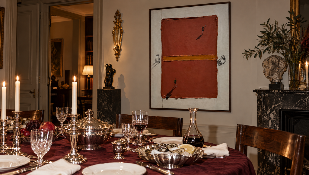 A framed abstract painting with a terracotta red and brown background hangs on a wall in a styled dining room. The room features a dining table set with silverware, wine glasses, and lit candles, with a fireplace and decorative plants in the background.