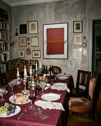 A styled dining room table set for a dinner party with a burgundy tablecloth, white plates, wine glasses, silverware, candles, and a teapot, surrounded by wooden chairs in a rustic room with a textured gray wall and framed art.