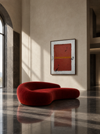 A modern, curved red velvet lounge chair in a minimalist, sunlit interior with a beige plaster wall, dark marble floor, and a framed abstract painting hanging above.