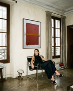 A woman in a black sleeveless dress sits in a black metal chair in a stylish room, with a large framed abstract painting on the wall behind her.