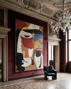 A large framed abstract painting with bold black, red, blue, cream, and tan shapes hangs on a deep burgundy wall in an ornate interior with a chandelier, carved ceiling, and black leather armchair.