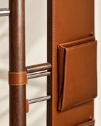 Close-up of a brown leather wall-mounted coat rack with a vertical pocket and metal bars, shown against a light neutral background.