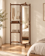 A tall freestanding wooden floor mirror with built-in shelves and hanging bars, styled in a warm, sunlit bedroom. The mirror reflects a bed in the background, and the product is shown in a real interior setting.