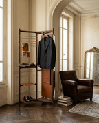 A tall dark wood coat rack with integrated shelves, a hanger bar, and a small mirror stands in a classic room with herringbone wood floors, next to a brown leather armchair.