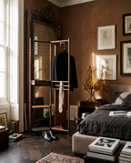 A tall, narrow wooden clothing rack with a mirror and open shelving stands in a styled bedroom corner. A dark jacket and tie hang on the rack against warm brown walls, with framed art, a bed, and a window in the background.