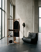 A modern interior scene with a black coat hanging on a tall wooden rack, a black leather lounge chair, a sculptural black floor lamp, and a small brown bag on a dark stone floor against a textured concrete wall and large windows.