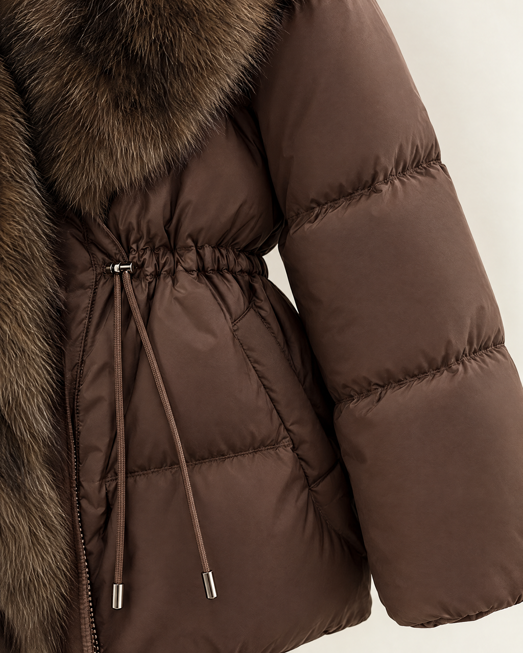 Close-up of a brown puffer jacket with a large brown fur collar and a drawstring waist, shown against a light neutral background.
