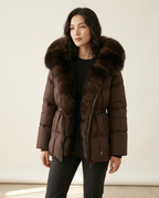 A woman wearing a dark brown puffer jacket with a large dark brown fur-trimmed collar, styled with a black top and black pants against a plain off-white studio background.