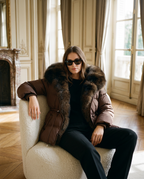 A woman wearing a brown puffer jacket with a large dark fur collar sits in a cream boucle armchair in an elegant room with wood floors and tall windows.